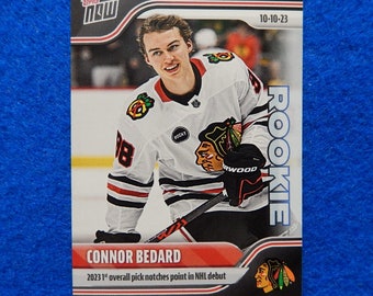Pode incluir: Um cartão de hóquei de Connor Bedard, a primeira escolha geral de 2023, em sua estreia na NHL. O cartão é vermelho, preto e branco com o texto "Rookie" e "10-10-23".