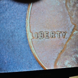 May include: Close-up of a copper-colored penny coin. The word "LIBERTY" is embossed on the coin's surface. The coin has a worn appearance, with visible details of the design.