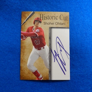 May include: A baseball card featuring a photo of Shohei Ohtani in a red baseball uniform, swinging a bat. The card has the text "Historic Cut" and "Shohei Ohtani" printed on it. The card also includes a purple signature.