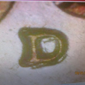 May include: A dark olive green letter D with a slightly distressed, textured appearance. The letter has a rounded, blocky shape with a lighter, off-white center. The date 10/31/2023 is printed in small, orange-brown text in the bottom right corner.