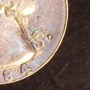 May include: Close-up of a silver coin with the partial image of a woman's head and the number "84" visible. The coin has a worn appearance, with details slightly blurred.