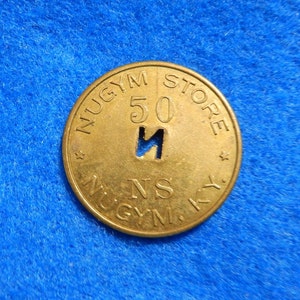 May include: A gold-colored metal token with the text "NUGYM STORE" and "50" printed around the edge. The center of the token has a large "N" with a hole cut out of it. The text "N.S. MUGYN." is printed below the "N".