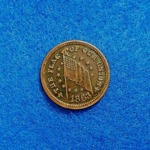 May include: A round copper-toned token featuring an embossed American flag at the center, surrounded by the text THE FLAG OF OUR UNION and the year 1863. The design includes a circular border of small stars encircling the central flag motif.