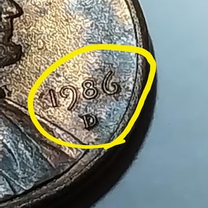 May include: A copper-colored United States one-cent coin featuring the year 1986 and a D mint mark stamped on the surface. The coin has a textured, metallic appearance with a yellow circular outline drawn around the date and mint mark area.