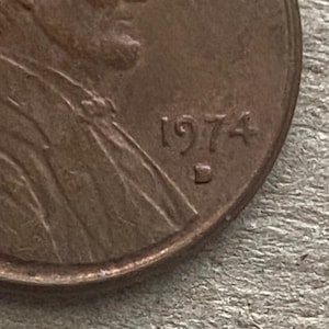 May include: Close-up of a copper-colored penny. The year "1974" is visible, along with the letter "D" below the date. The coin's surface shows signs of wear and age, with details of a portrait on the left side.
