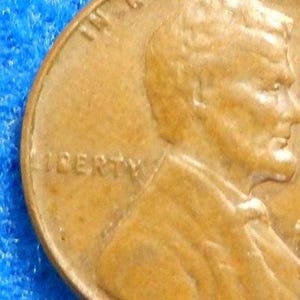 May include: Close-up of a copper-colored coin with the word "LIBERTY" visible. The profile of a person is embossed on the coin. The coin is set against a blue background.