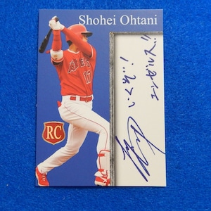 May include: A baseball card featuring Shohei Ohtani, a baseball player for the Los Angeles Angels. The card is blue with a red, white, and blue baseball uniform. The card has a gold border and a red and gold "RC" logo. The card is signed by Shohei Ohtani in blue ink.