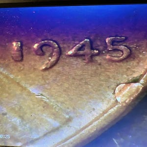 May include: Close-up of a copper-colored coin with the year 1945 visible. The numbers are raised and slightly worn, with a small, irregular shape on the coin's edge. The background has a gradient of gold, brown, and blue.