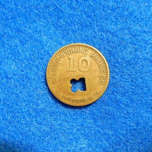 May include: A gold-colored metal token with the text "RALEIGH WYOMING MINING CO." and the number "10" in the center. The token has a hole in the shape of a cross.
