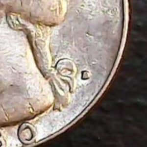 May include: Close-up of a silver coin, showing a partial image of a person and the letter "D". The coin has a worn appearance, with visible scratches and imperfections on its surface. The details of the design are partially visible.