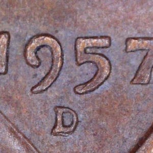 May include: A close-up view of a copper-colored metal surface featuring the embossed date 1957 and a capital letter D mint mark positioned directly below the year. The texture appears weathered with a brownish, metallic patina across the entire surface.