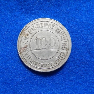 May include: A silver coin with the text "HARLAN RIDGEWAY MINING CO. KYO" around the edge. The number "100" is in the center. The coin is on a blue background.