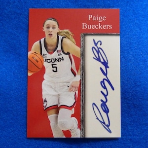 May include: A red and white basketball card featuring Paige Bueckers, a basketball player wearing a white jersey with the text "UCONN 5" and a basketball in her hands. The card has a silver border and a white signature box with the text "Bueckers" in blue ink.