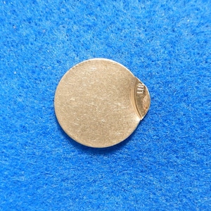 May include: A silver-colored round metal disc with a slightly damaged edge. The disc has a faint inscription on the edge that reads "UR".