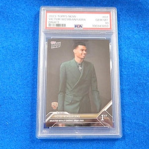 May include: A 2023 Topps Now Victor Wembanyama Draft trading card graded PSA Gem Mt 10. The card features a photograph of Victor Wembanyama wearing a dark green double-breasted suit, with text including 1st Pick and Selected with 1st Overall Draft Pick.