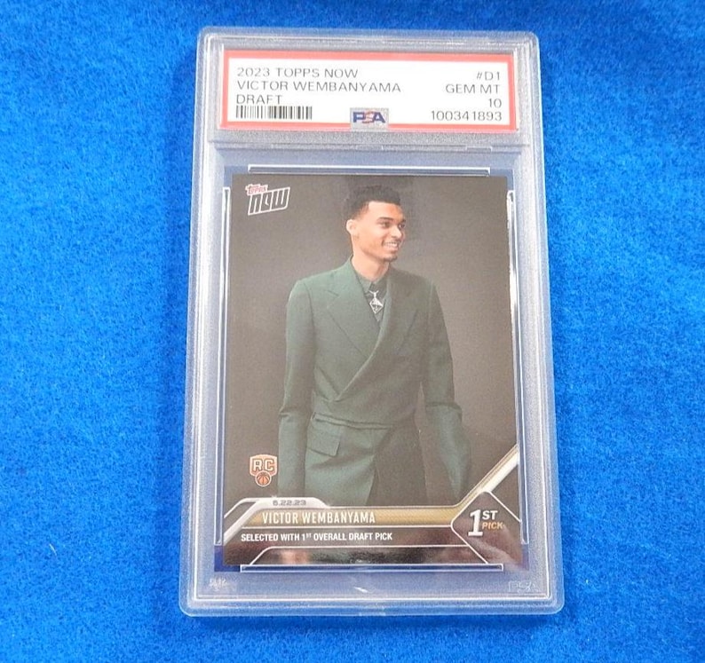 May include: A 2023 Topps Now Victor Wembanyama Draft trading card graded PSA Gem Mt 10. The card features a photograph of Victor Wembanyama wearing a dark green double-breasted suit, with text including 1st Pick and Selected with 1st Overall Draft Pick.
