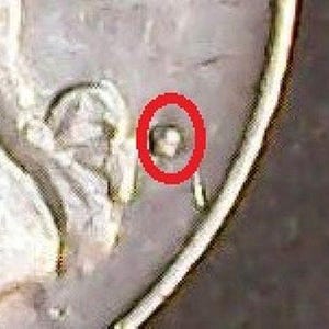 May include: A metallic coin surface featuring a raised, textured detail highlighted by a bright red circular outline. The coin exhibits a weathered, silver-toned finish with visible surface imperfections and metallic contours surrounding the marked area.