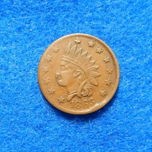 May include: A copper-colored 1863 Indian Head penny. The coin features a profile of a Native American in a feathered headdress, surrounded by stars. The year 1863 is visible below the profile. The coin is set against a blue background.