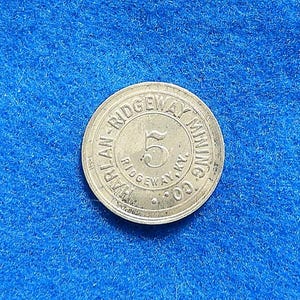 May include: A circular metallic token featuring the text HARLAN RIDGEWAY MINING CO. and RIDGEWAY, KY. with the number 5 prominently displayed in the center. The token has a raised border and a smooth, reflective surface finish.
