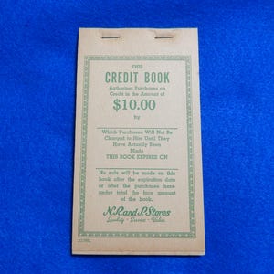 May include: A vintage brown credit book with green text that reads "This Credit Book Authorizes Purchases on Credit in the Amount of $10.00 by Which Purchases Will Not Be Charged to Him Until They Have Actually Been Made This Book Expires On No safe will be made on this book after the expiration date or after the purchases here under total the face amount of the book. N.Rand P.Stores Quality Service Value"