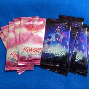 May include: Several packets of McDonald's breakfast meal and HuntraX meal. The breakfast meal packets are pink and white with "The Sajia Boys" text. The HuntraX meal packets are dark blue with a silhouette design and "The HuntraX Meal" text.
