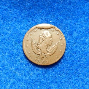 May include: A copper-colored coin with a profile of a man on one side and a design of two flags with stars on the other side. The year 1862 is inscribed on the coin.