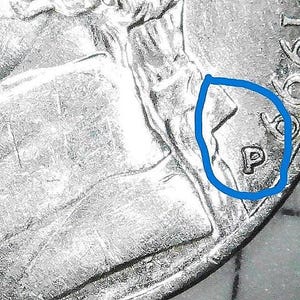 May include: A close-up view of a silver-coloured coin featuring a raised letter P mint mark circled in blue ink. The coin surface shows metallic textures and relief details of a portrait, with the partial date 199 visible in the upper right corner.