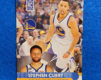 Könnte beinhalten: Eine Rookie-Sammelkarte mit Stephen Curry von den Golden State Warriors. Die Karte ist blau und gelb mit einem Foto von Curry in seiner Uniform. Auf der Karte steht "Rookie RC Card" und "Golden State Warriors".