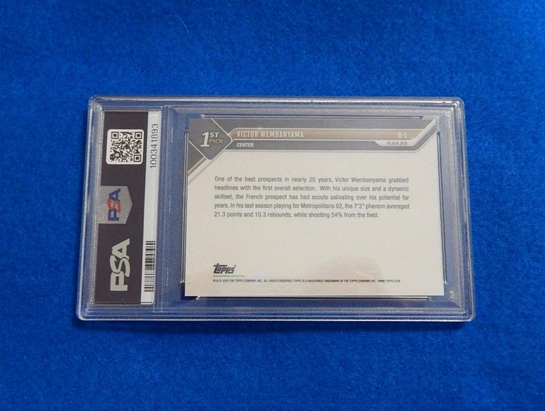 May include: A PSA-graded Victor Wembanyama Topps trading card encased in a clear plastic holder. The back of the card features white text on a light background, detailing the player's stats and history, with a QR code and PSA logo on the left side.
