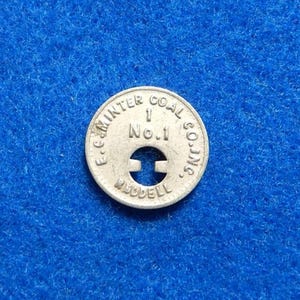 May include: A silver-colored metal token with the text "E.C. MINTER COAL CO. INC." around the edge. The token has a central cutout and the number "1 No.1" in the middle. The token is on a blue background.