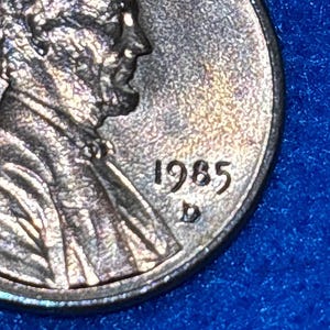 May include: A metallic copper-colored United States one-cent coin featuring the profile of Abraham Lincoln facing right. The date 1985 is clearly visible to the right of the portrait, with a small D mint mark located directly below the year.