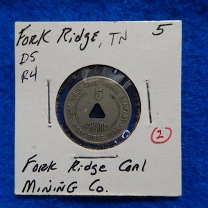 May include: A silver token with the text "Fork Ridge Coal Mining Company" and "Fork Ridge, Tenn." around the edge. The center of the token has a triangle with the number "5" above it. The token is on a white card with the text "Fork Ridge, TN 5" and "Fork Ridge Coal Mining Co."