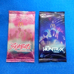 May include: Two McDonald's meal wrappers. One is pink with the text "The Saja Boys Breakfast Meal" and the McDonald's logo. The other is dark blue with the text "The Huntrx Meal" and the McDonald's logo.