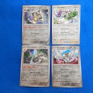 May include: Four Pokémon trading cards on a blue surface. The cards feature Aegislash, Tornadus, Ferrothorn, and Doublade. Each card has a unique illustration and text describing abilities and stats. The cards are in English.