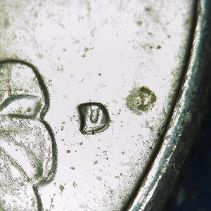 May include: A close-up view of a metallic coin surface featuring a stamped letter D mint mark next to a small, circular, textured indentation. The surface shows signs of wear, including fine scratches and small pits across the silver-toned metal.