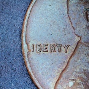 May include: Close-up of a copper-colored penny, showing the word "LIBERTY" in raised letters. The coin's surface has a textured appearance, with a portion of a profile visible. The image is set against a blue background.