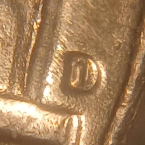 May include: A close-up view of a metallic surface featuring a stamped letter D. The texture appears raised and embossed, showing a copper-toned finish with visible metallic grain and light reflections across the surface of the coin.