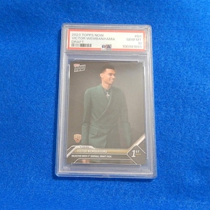 May include: A 2023 Topps Now Victor Wembanyama Draft trading card encased in a PSA slab. The card features a portrait of Victor Wembanyama in a dark green blazer, with text reading Topps Now, Victor Wembanyama, and Selected with 1st Overall Draft Pick.