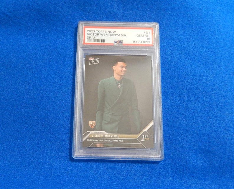 May include: A 2023 Topps Now Victor Wembanyama Draft trading card encased in a PSA slab. The card features a portrait of Victor Wembanyama in a dark green blazer, with text reading Topps Now, Victor Wembanyama, and Selected with 1st Overall Draft Pick.