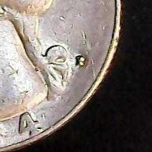 May include: Close-up of a silver coin with the letters "A" and "D" visible. The coin has a worn appearance with visible scratches and imperfections. The edge of the coin is gold-colored.