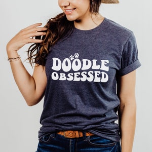 Op de afbeelding: Een marineblauw T-shirt met witte tekst die "Doodle Obsessed" luidt in een retro-lettertype. De tekst wordt vergezeld door drie witte potafdrukken.