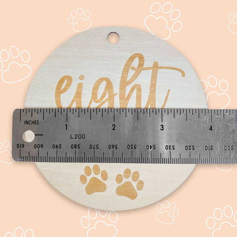 May include: A round wooden ornament with the word "eight" engraved in a cursive font. The ornament is approximately 4 inches in diameter and has two paw prints at the bottom.