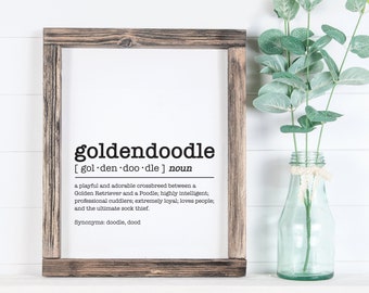Puede incluir: Una impresión enmarcada con fondo blanco y texto negro que define la palabra "goldendoodle" como un cruce juguetón y adorable entre un Golden Retriever y un caniche. El texto los describe como altamente inteligentes, abrazadores profesionales, extremadamente leales y amantes de la gente. También afirma que son el ladrón de calcetines definitivo. La impresión enumera "doodle" y "dood" como sinónimos.