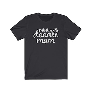 Op de afbeelding: Donkergrijs t-shirt met de witte tekst "mini doodle mom" in sierlijke letters, vergezeld van twee kleine pootafdrukken. Het shirt heeft korte mouwen.