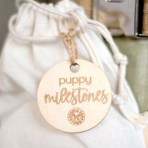 May include: A round wooden tag with the text "puppy milestones" in cursive script. The tag is attached to a white drawstring bag.