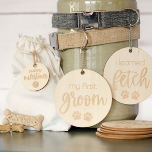 May include: A set of three wooden dog tags with paw prints and text. The tags read "puppy milestones", "my first groom", and "i learned fetch".