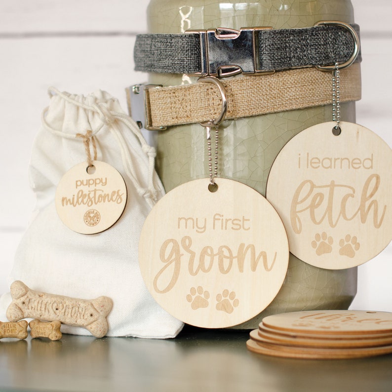 May include: A set of three wooden dog tags with paw prints and text. The tags read "puppy milestones", "my first groom", and "i learned fetch".