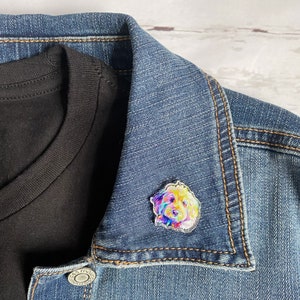 May include: A colorful dog-themed pin is attached to a denim jacket. The pin features a vibrant watercolor-style portrait of a dog with yellow, purple, blue, and pink hues. A black t-shirt is visible beneath the jacket.