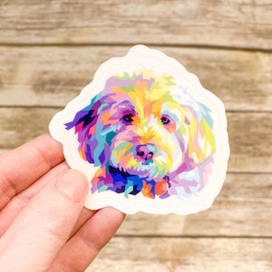 May include: A colorful sticker featuring a stylized portrait of a dog. The dog's face is rendered in a mosaic of vibrant colors, including yellow, purple, blue, and pink. The sticker has a white border and a unique shape.