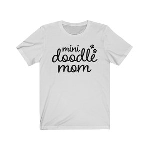 Op de afbeelding: Wit T-shirt met zwarte tekst die "mini doodle mom" luidt met drie poot afdrukken boven het woord "doodle".